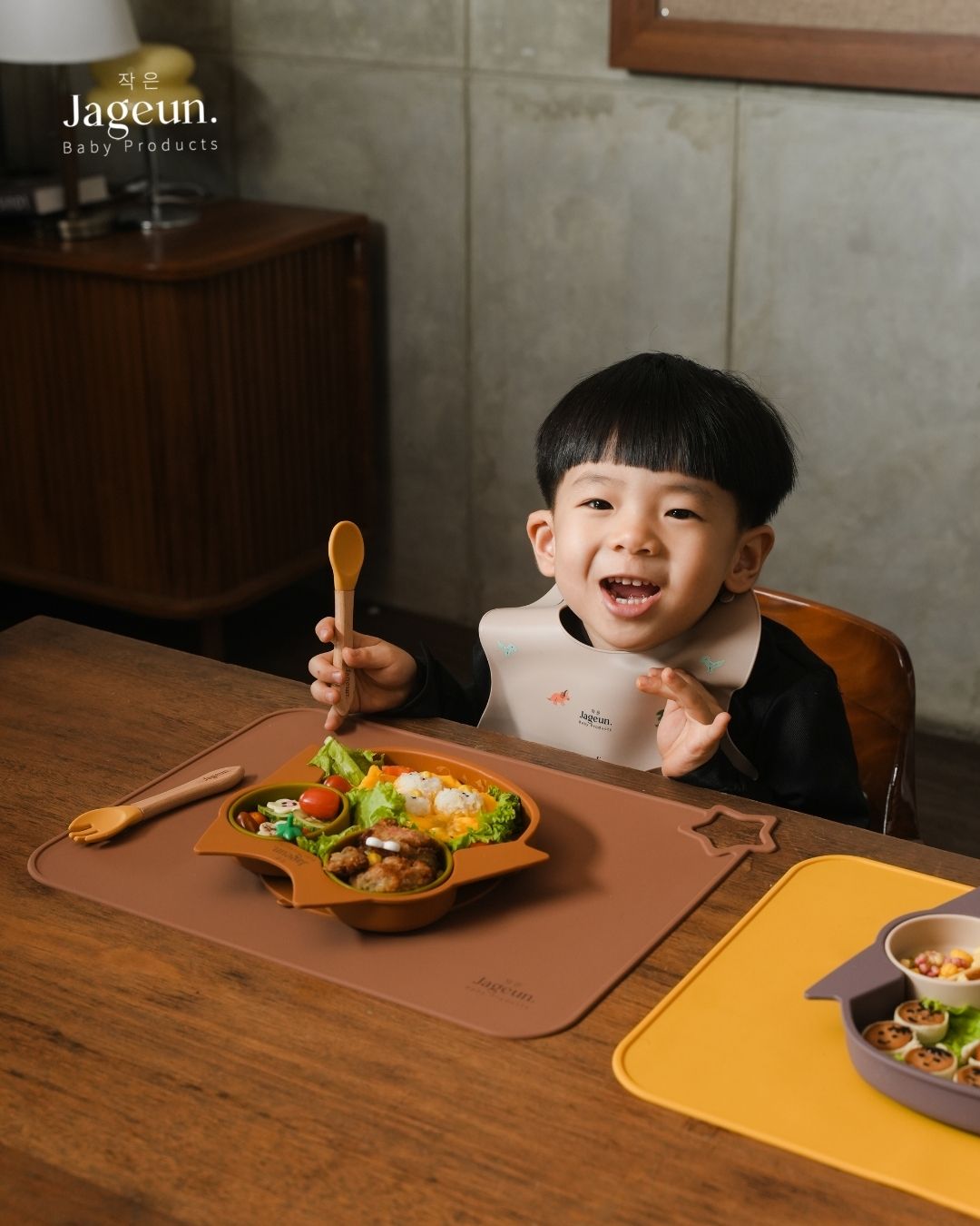 Anak kecil tersenyum sambil memegang sendok Jageun di meja makan